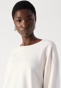 Lichtbeige sweatshirt met een relaxte pasvorm en brede halslijn. Bevat een geborduurd logo op de borst en een zachte, gestructureerde stof.