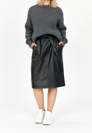 Femme portant un gros pull en maille grise rentré dans une jupe midi en cuir noir avec une taille nouée et des baskets blanches.