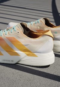 Een paar witte en gele hardloopschoenen met lichtblauwe veters die rusten op een gestructureerd buitenshuis oppervlak in zonlicht.
