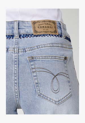 Hellblaue Jeans aus Denim mit einem Logo-Etikett aus braunem Leder. Sie sind mit dekorativer roter Stickerei am Taillenbund und einer Tasche mit geschwungener Stickerei versehen.