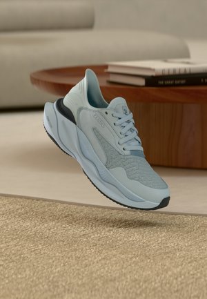 Lichtblauwe sportschoen met een textuurlzool en veters, zwevend boven een geweven beige vloerkleed in een moderne woonkameromgeving.