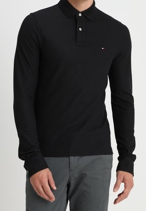 Mann trägt ein schwarzes Langarm-Poloshirt mit weißen Knöpfen und graue Hose vor einfachem Hintergrund.