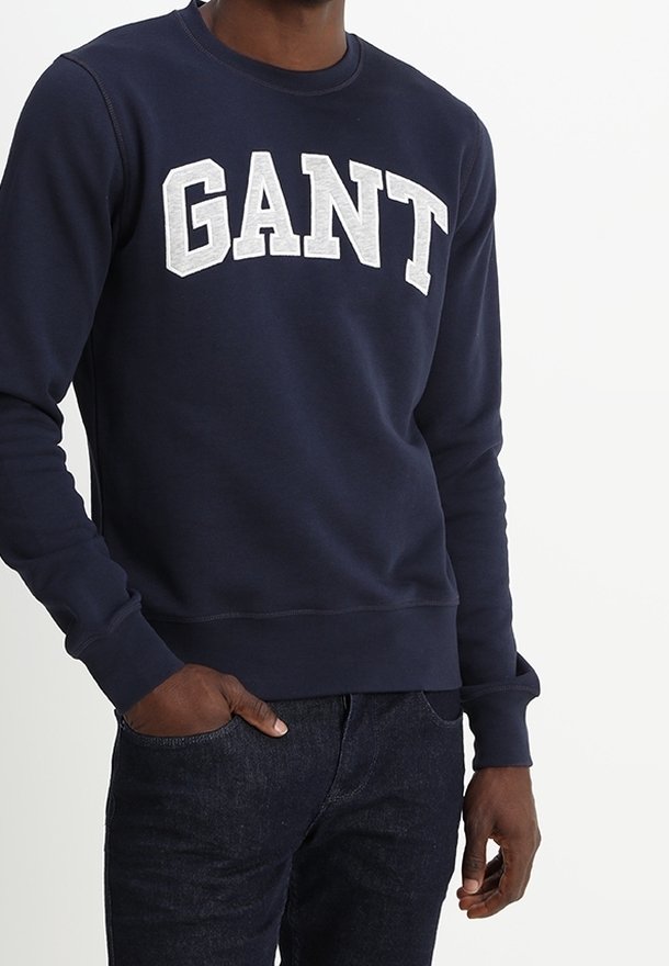 Homme portant un sweat-shirt bleu marine avec "GANT" en grandes lettres blanches sur la poitrine et un jean bleu foncé.