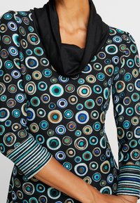 Czarny top z dekoltem w kształcie cowl, z wzorem kolorowych koncentrycznych kółek w odcieniach turkusu, niebieskiego i brązowego. Paski przy mankietach dodają szczegółów.