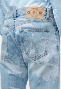 Světle modré džínsy s batikovým vzorem připomínajícím vybledlé mraky, viditelnými zadními kapsami a béžovou koženou nášivkou s nápisem Fiorucci na pásku.