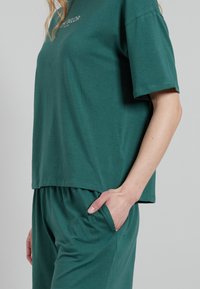 Green cotton t-shirt with a relaxed fit, short sleeves, and a rounded hem. Features a side pocket on matching pants.