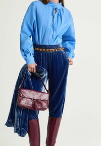 Blusa azzurro chiaro con dettaglio collo annodato, pantaloni in velluto blu navy con vita arricciata, borsa in pelle bordeaux e stivali abbinati al ginocchio.