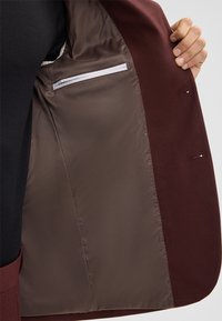 Interno di giacca in tessuto bordeaux con fodera liscia marrone; presenta una tasca con zip e vestibilità su misura. Dettagli in tessuto testurizzato visibili.