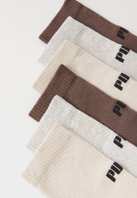 Conjunto de cinco pares de calcetines en tonos marrón, beige y gris, con puños acanalados y un tejido texturizado, presentando el logotipo negro en el lateral.