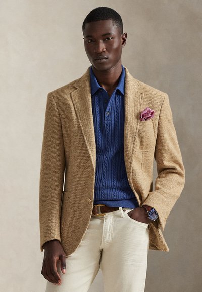 Beżowa marynarka w jodełkę na niebieskim swetrze polo o splocie warkocza, zestawiona z jasnymi spodniami. Różowa poszetka w paisley i zegarek dopełniają całość.