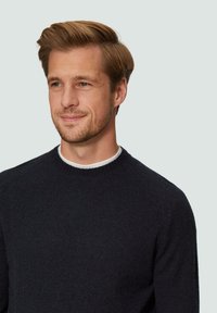 Donkere gebreide trui met een ronde hals, geribde manchetten en rechte zoom. Draagster glimlacht, met een lichte achtergrond.