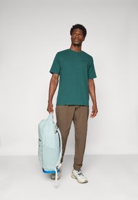 Bărbat îmbrăcat cu tricou verde și pantaloni maronii, ținând un rucsac bleu deschis, pe un fond alb simplu.