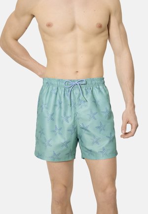 Hombre con bañador azul claro con patrón de estrellas de mar en azul oscuro y cintura con cordón, de pie frente a un fondo liso.