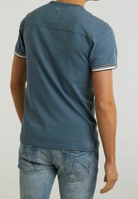 Muž v modré košili s krátkým rukávem a bílými proužky na manžetách, ve vybledlých modrých džínách, stojí zády k dějiště před jednobarevným pozadím.