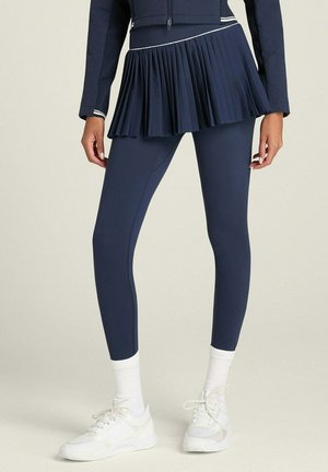 Femme portant une jupe plissée bleu marine sur des leggings assortis, des chaussettes blanches et des baskets blanches, debout devant un fond clair uni.
