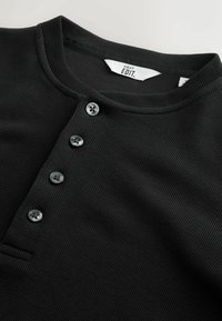 Schwarzes, geripptes Henley-Shirt mit rundem Halsausschnitt, fünf grauen Knöpfen und einem kleinen Taschen-Detail. Stoff aus Baumwollmischung, strukturierte Oberfläche.