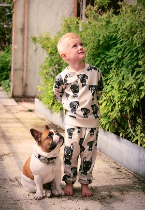 Helles beigefarbenes Bekleidungsset mit Hundeprints; langärmeliges Oberteil und Hose. In der Nähe sitzt ein braun-weißer Französischer Bulldogge. Grüne Pflanzen im Hintergrund.