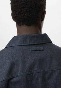 Giacca in denim scuro, con colletto classico e dettaglio sul yoke posteriore. Il tessuto ha un aspetto strutturato e una leggera variazione di colore.
