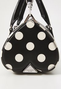 Heart-shaped black leather handbag with large white polka dots, featuring a silver triangular emblem and dual top handles.