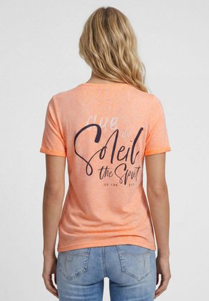 Blonde woman facing away wearing an orange t-shirt with "Club du Soleil the Spirit of the Sun" text and light blue jeans.