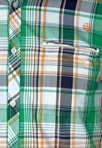 Camisa de manga corta a cuadros en verde, blanco, naranja y azul marino. Cuenta con un bolsillo en el pecho con cremallera y botones blancos. Tela con textura suave.