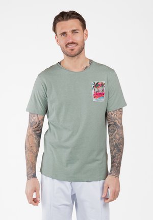 Mann mit Tätowierungen an den Armen, der ein hellgrünes T-Shirt mit Palmenmotiv und weiße Hosen trägt, steht vor einem weißen Hintergrund.
