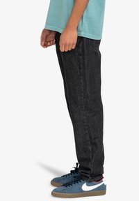 Jeans de mezclilla negra con corte recto, combinados con zapatillas azules que tienen detalles en blanco. La tela tiene una textura sutil y detalles de costura mínimos.
