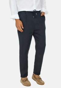 Pantaloni di lino navy con vita regolabile e davanti plissettato. Gambe aderenti e affusolate, in tessuto leggero, abbinati a mocassini color cammello.