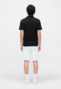 Férfi áll háttal, fekete, rövid ujjú ingben, amelyen függőleges szövegminta látható, fehér térdig érő rövidnadrágban, fehér zokniban és fehér tornacipőben.