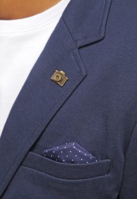 Marinblå blazer med en guldkamera nål på slaget och en marinblå fickruta med vita prickar i bröstfickan.