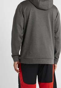 Haut à capuche gris avec une texture lisse, doté d'une grande capuche et de longues manches. Associé à un short de sport noir et rouge.