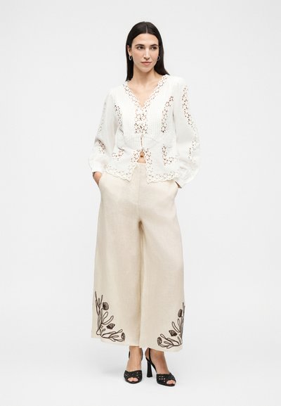 Γυναίκα που φοράει λευκή δαντελωτή μπλούζα, μπεζ φαρδιά παντελόνια με μαύρη κεντητή λουλουδάτη διακόσμηση κοντά στο τελείωμα και μαύρα ψηλοτάκουνα σανδάλια, στέκεται με τα χέρια στις τσέπες.