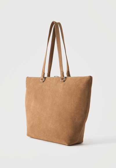 Sac cabas en daim marron avec deux longues bandoulières et une fermeture éclair sur le dessus, présenté sur un fond clair uni.