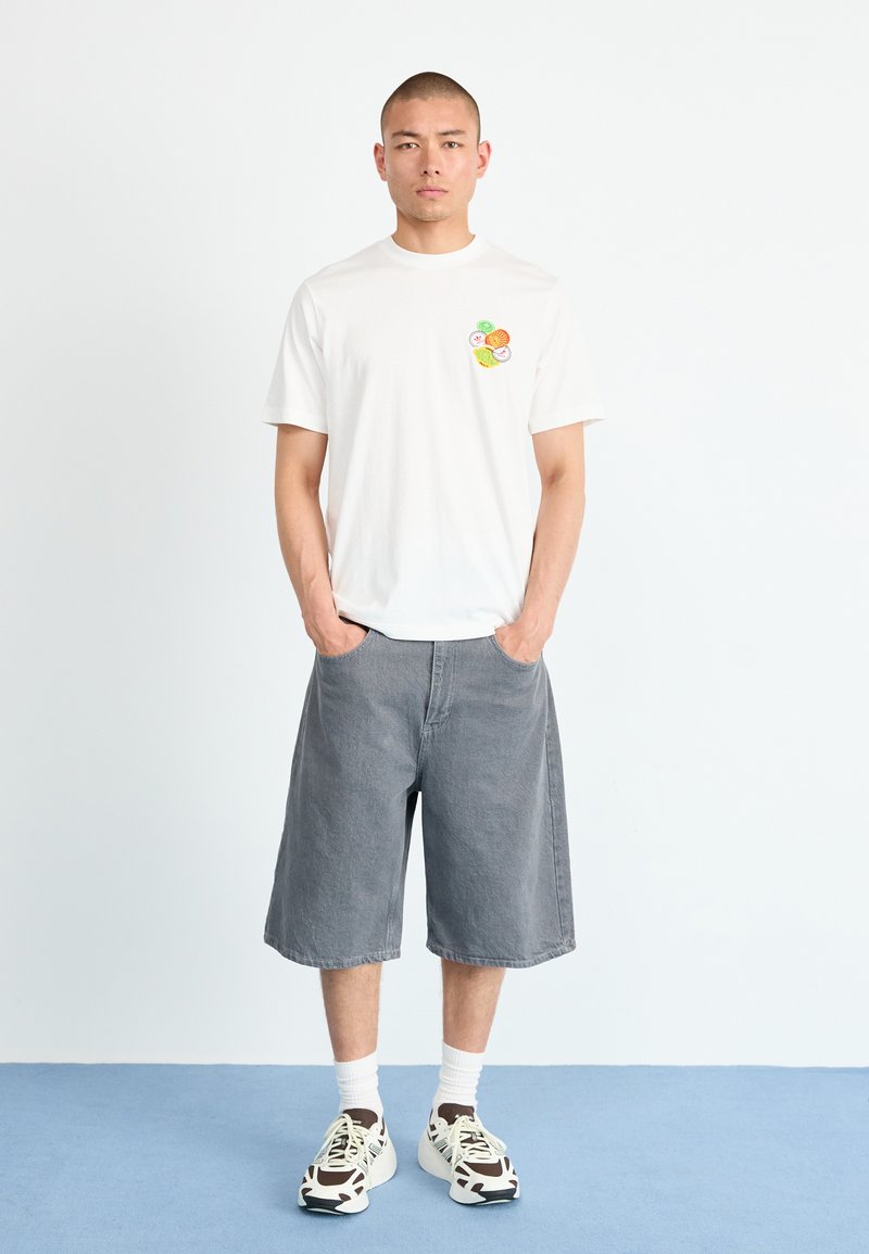 Weißes T-Shirt mit kurzen Ärmeln und bunten Obstgrafiken auf der Brust, kombiniert mit lockeren grauen Jeansshorts und klobigen Sneakers.