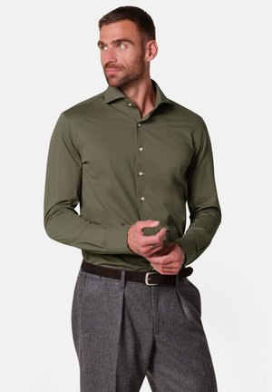 Mand iført olivengrøn knap-til-shirt og grå bukser, der justerer sin venstre manchet, kigger mod venstre på en ensfarvet hvid baggrund.