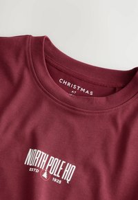 Burgundské tričko s okrúhlym výstrihom vyrobené z bavlny, s bielym grafickým textom: "NORTH POLE HQ ESTD 1925," a ikonou stromy.