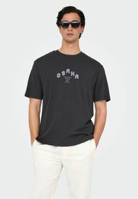 Mørkeblå bomulds t-shirt med "OSAKA" grafisk tryk i hvid og lilla. Rund hals, korte ærmer. Båret med lyse bukser.