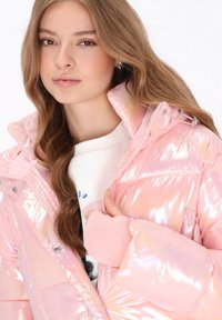 Kobieta o długich brązowych włosach, ubrana w błyszczącą różową kurtkę puchową i biały top, trzymająca kołnierz kurtki jedną ręką.