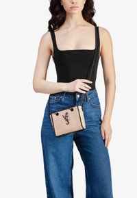 Donna che indossa un top senza maniche nero e jeans blu, mentre tiene in mano una borsa a tracolla beige intrecciata con bordi neri e tracolla a catena.
