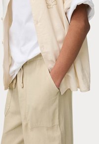 Chemise en lin beige à manches courtes et fermeture à boutons, portée par-dessus un t-shirt blanc. Pantalons beiges clairs à cordon de serrage avec poches latérales.