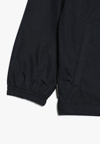 Manica di giacca nera con polsino elastico, dotata di tasca con zip laterale e texture liscia. Logo ricamato vicino all'orlo.