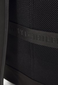 Zaino nero con un pannello in rete, texture liscia e una prominente tracolla nera con dettagli del logo in rilievo. Design moderno per versatilità.