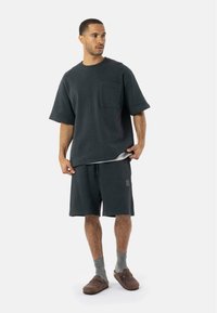 Sweatshirt ample gris foncé à manches courtes avec une poche frontale, associé à un short assorti. Le mannequin porte des chaussettes et des chaussures à enfiler marron.