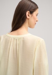 Camicia gialla chiaro con una texture morbida e trasparente, caratterizzata da un collo a barca e dettagli arricciati sul retro.