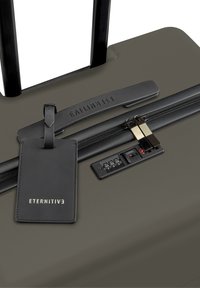 Valigia verde scuro con guscio rigido, manico nero estraibile, meccanismo di chiusura a zip combinato con lucchetto e un'etichetta in pelle nera con scritto "ETERNITIV."