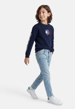 Dreng, der går i lyseblå jeans, hvide sneakers og marineblå sweatshirt med teksten 'peace and love' på ærmet og farverigt emblem på brystet.