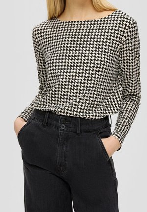 Femme portant un haut à manches longues noir et blanc à motif pied-de-poule, rentré dans un jean noir taille haute, les mains dans les poches.
