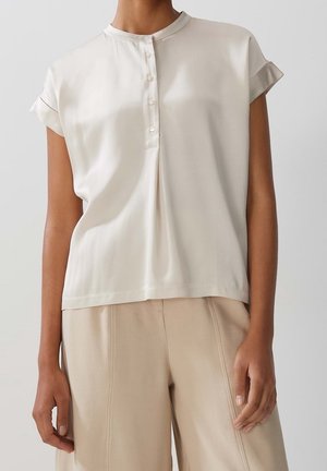 Femme portant un chemisier ivoire à manches courtes et boutonné, associé à un pantalon beige taille haute à jambes larges, sur un fond uni.