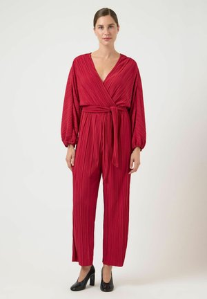 Röd plisserad jumpsuit med djup V-ringning och vida ärmar, knuten i midjan. Bärs med svarta klackskor med en slät yta.