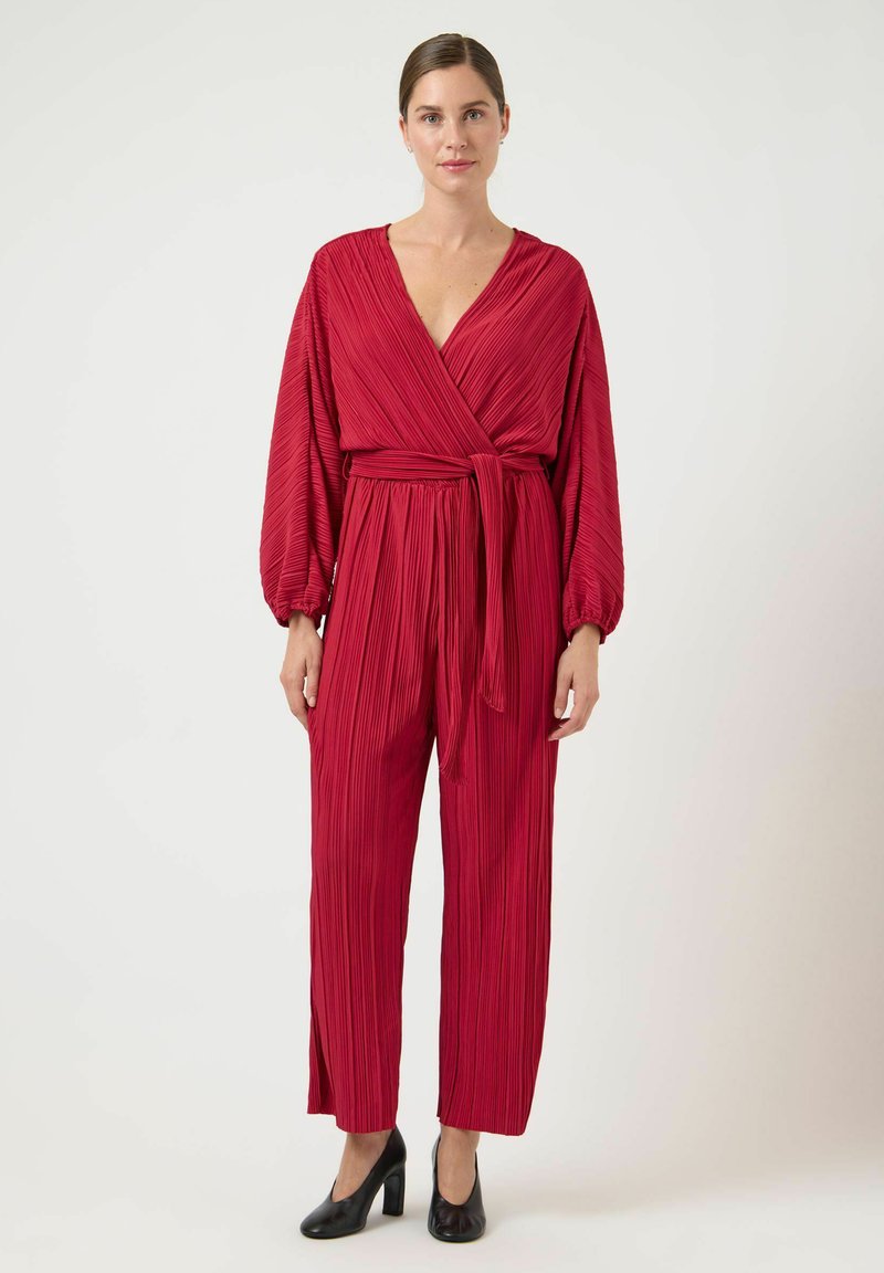 Rød plisseret jumpsuit med dyb V-udskæring og brede ærmer, bundet om taljen. Båret med sorte høje sko med en glat tekstur.